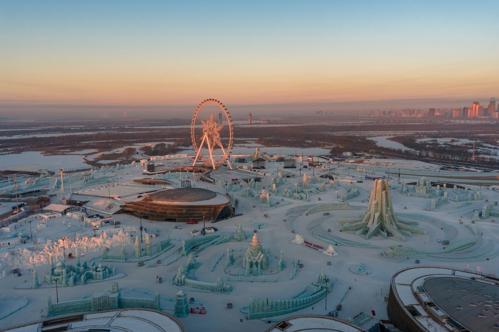 Aerial landscape photograph taken with the DJI Mavic 4 Pro, capturing intricate ice sculptures and a giant Ferris wheel at sunrise in a snow-covered winter theme park — showcasing exceptional dynamic range and colour depth.